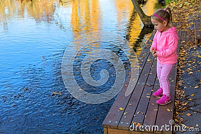 Adorable happy girl feeding fish in autumn park. Young female throwing bread in to the lake to feed fish. Stock Photo