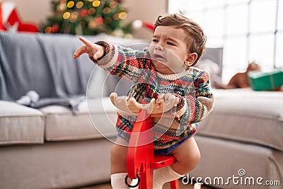 Adorable hispanic toddler crying and playing with reindeer rocking by christmas tree at home Stock Photo