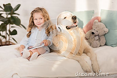 adorable kid holding tablet, funny golden retriever with headphones lying on bed Stock Photo