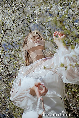 Adorable lady in wedding gown with diaphanous sleeves close eyes and enjoy sun while standing in white cherry blossoms. Stock Photo