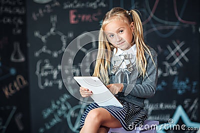 Adorable little blond schoolgirl in school uniform holding white digital tablet. Chalkboard with school formulas background. Back Stock Photo