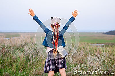Adorable little girl with long braided hair in plaid skirt on hill with raised hands up to the sky. Happy traveler child Stock Photo