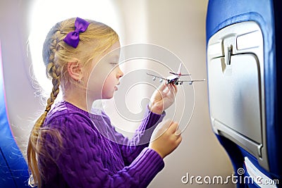 Adorable little girl traveling by an airplane. Child sitting by aircraft window and playing with toy plane. Traveling with kids. Stock Photo