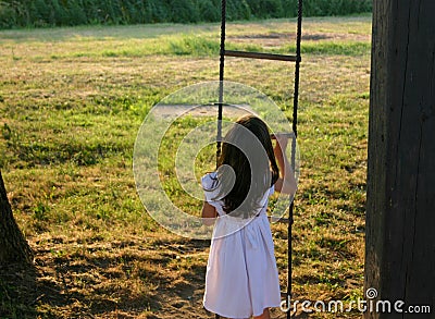 Adorable little girl in a white dress holding from rope stairs in a garden captured from behind Stock Photo