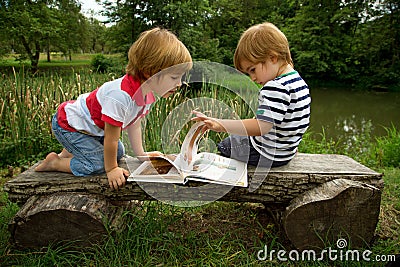 Adorable Little Twin Brothers Sitting on a Wooden Bench and Looking at Interesting Pictures in the Book Near the Beautiful Lake Stock Photo