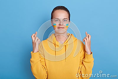 Adorable positive hopeful woman wearing hoodie with blue and yellow flag on cheeks, standing isolated over blue background and Stock Photo