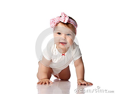 Adorable positive little baby girl in white body, hair bow decoration and barefoot creeping on floor, smiling and looking aside Stock Photo