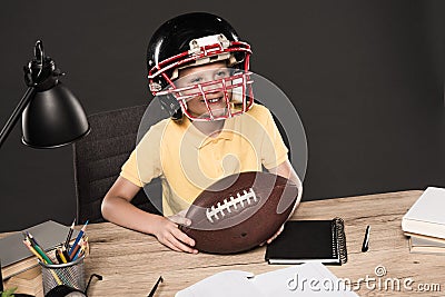 adorable schoolboy in american football helmet sitting at table with ball books lamp colour pencils textbook on grey Stock Photo