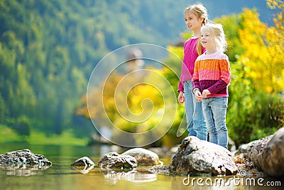 Adorable sisters playing by Konigssee lake in Germany on warm summer day. Cute children having fun splashing water and throw Stock Photo