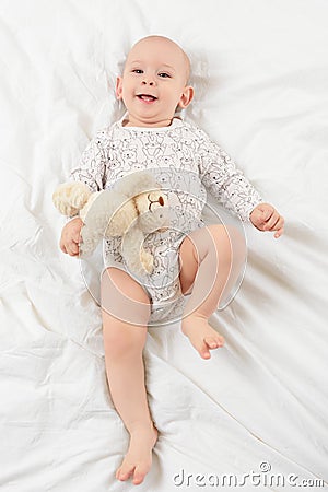 Adorable smiling baby boy with blue eyes lying on a bed with his favourite stuffed teddy bear toy, looking at camera. Stock Photo