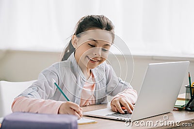 Smiling child writing in copy book and using laptop while doing homework Stock Photo