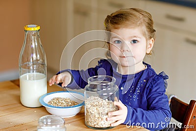 Adorable toddler girl eating healthy oatmeals with milk for breakfast. Cute happy baby child in colorful clothes sitting Stock Photo