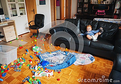 Adorable 1 year old baby boy with funny facial expression playing in a very messy living room Stock Photo