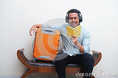 Adult Asian man sitting with travelling suitcase beside while listening music and read a book Stock Photo