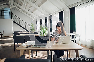 Adult attractive woman checking phone while sitting with a laptop at the table in a cafe. Stock Photo