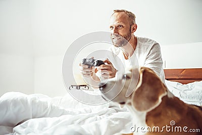 Adult breaded man waked up and plays PC games don`t stands up from bed.His beagle dog watching the game with very interest Stock Photo