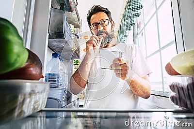 Adult caucasian man taking note list of food looking inside the open fridge at home - kitchen activity and alternative point of Stock Photo