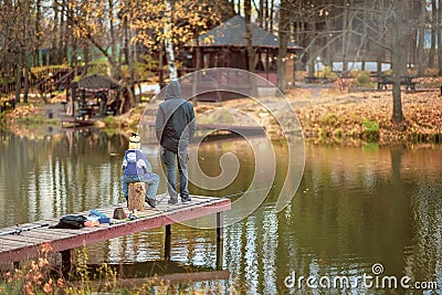 Adult and child on a wooden pier, fishing bridge. Autumn park. Fallen leaves, sunny day, natural background. Stock Photo