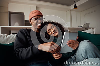 Trendy adult couple, multi-ethnic, laughing at video on tablet on sofa in modern apartment Stock Photo