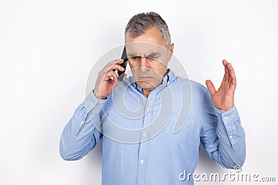 Adult handsome man with grey hair wearing blue shirt having phone conversation looks desperate standing on white Stock Photo