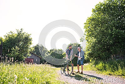 An adult hipster son with bicycle and senior father walking in sunny nature. Stock Photo