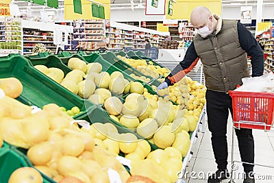 Adult man choosing fruit at a supermarket with gloves and a mask on. Bright red shopping cart next to him. Conscientious shopping Stock Photo