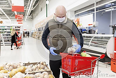 Adult man choosing vegetables at a supermarket with gloves and a mask on. Bright red shopping cart next to him. Conscientious Stock Photo