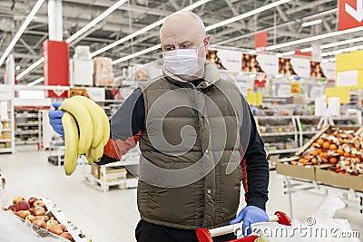 Adult man with gloves and a mask on holding bananas in the fruit aisle at a supermarket. Conscientious shopping during the virus Stock Photo