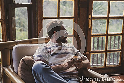 Adult mature man sitting on the chair drinking tea and looking outside the windows at home. Concept of people in indoor leisure Stock Photo