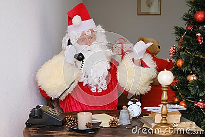 Adult santa claus in red suit sits at the table, calls on old phone, tree is beautifully decorated with balls, the concept of Stock Photo