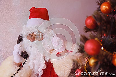 Adult santa claus in red suit sits at the table, calls on old phone, tree is beautifully decorated with balls, the concept of Stock Photo