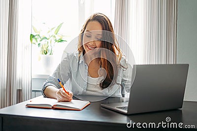 Adult smiling brunette woman doing notes in daily book with opened laptop. She study something with online course at the home Stock Photo