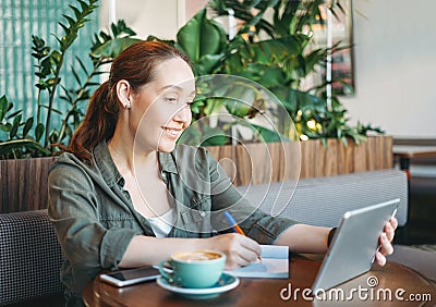 Adult smiling brunette woman doing notes and look at tablet takes online course on table at cafe Stock Photo