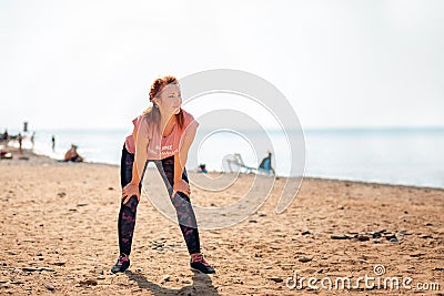 An adult tired beautiful woman in sportswear takes a break during a workout. In the background, the ocean and the beach. Copy Stock Photo