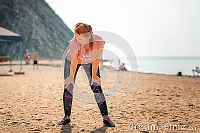 An adult tired caucasian woman in sportswear takes a break during a workout. In the background, the ocean and the beach Stock Photo