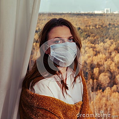 Adult woman in a medical mask by the window wrapped in a yellow plaid. Sad autumn weather and home comfort Stock Photo