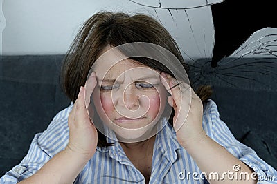Adult woman sits on a sofa, covered with a blanket, clasped her head in her hands, grimace of suffering on her face, the concept Stock Photo