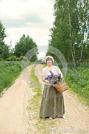 Adult woman walks through the woods park with a wicker basket with a bouquet of lupine flowers Stock Photo
