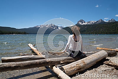 Adult woman 30-35 years sits on a pile of logs on the shoreline of Redfish Lake in Stanley Idaho in the Sawtooth Mountains in Stock Photo