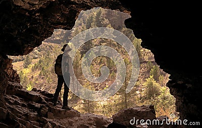 ADVENTURER WITH HAT AND BACKPACK IN A CAVE IN FRONT OF A PINE FOREST Stock Photo