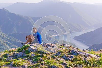 Adventurous man sitting on top of a mountain and enjoying the beautiful view, while looking downhill at the blue river and amazing Stock Photo