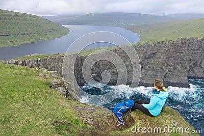 AERIAL: Unrecognizable woman sits on the edge of a cliff and looks at the lake. Stock Photo