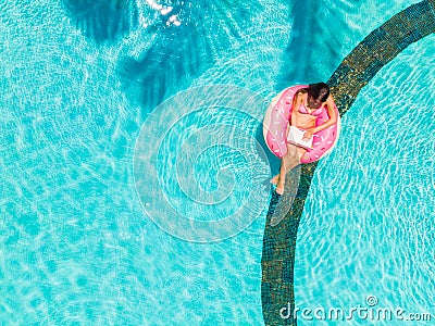 Aerial view of a young brunette woman swimming on an inflatable big donut with a laptop in a transparent turquoise pool. Stock Photo