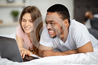 Affectionate multiracial couple spending time together at home, using laptop on bed Stock Photo