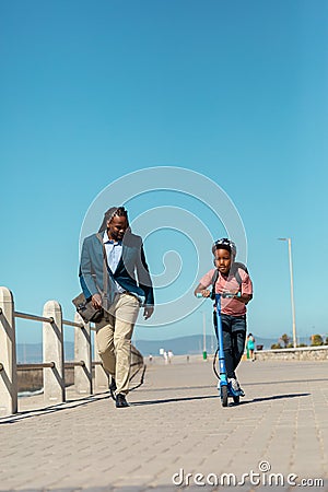 African american boy riding push scooter by father walking on promenade against clear blue sky Stock Photo