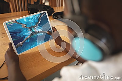 African american boy wearing headphones at home watching swimming competition on tablet Stock Photo