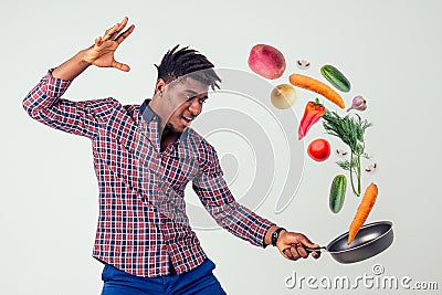 African american chef kitchener holding a frying pan wizard man cooking magic flying food salad, carrot, garlic, onion Stock Photo