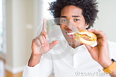 African American hungry man eating hamburger for lunch surprised with an idea or question pointing finger with happy face, number Stock Photo