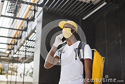 African American male courier food delivery in face mask with yellow thermal backpack Stock Photo