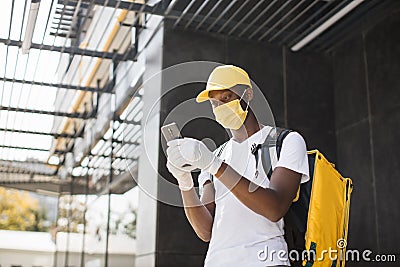 African American male courier food delivery in face mask with yellow thermal backpack Stock Photo